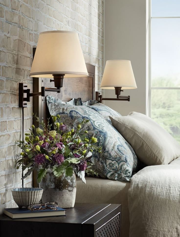 Bronze swing-arm lamp with an adjustable hinged arm and cone-shaped shade, mounted beside a bed and angled downward to provide focused reading light against a neutral wall.