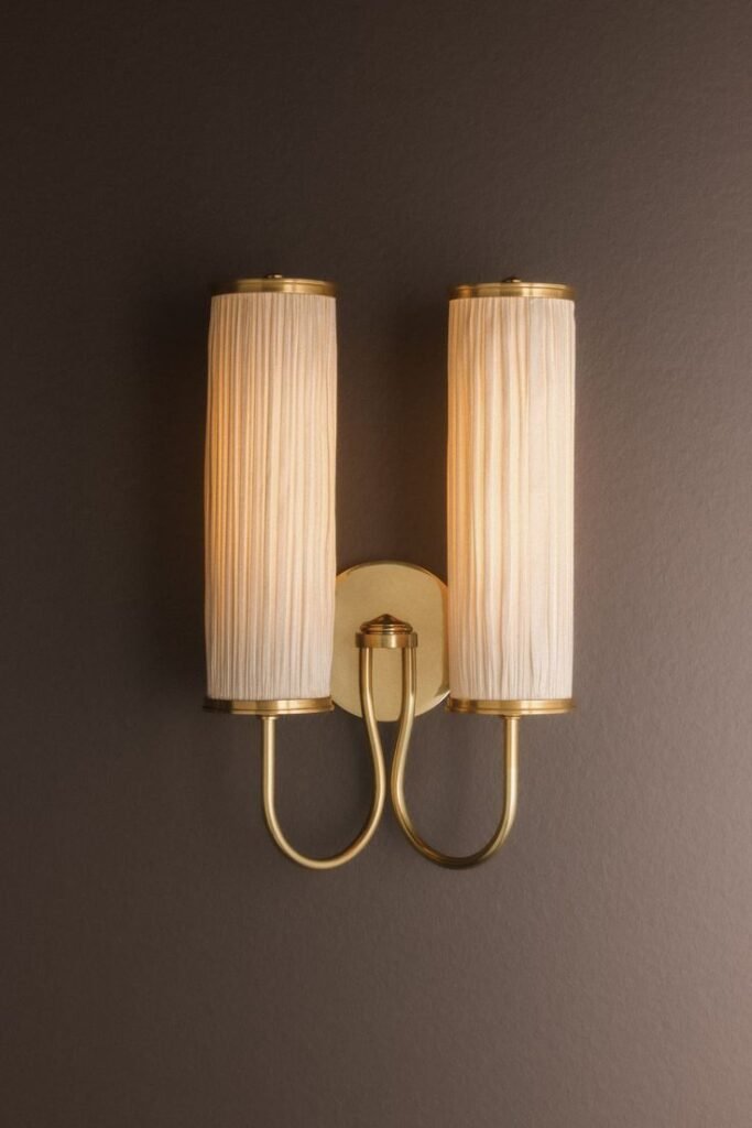 Elegant wall sconce with a soft, pleated white silk shade mounted on a polished brass arm, casting a warm, diffused glow against a black wall.